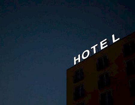 A building with a large illuminated "HOTEL" sign on the roof stands against a darkening sky, suggesting it's evening or nighttime.